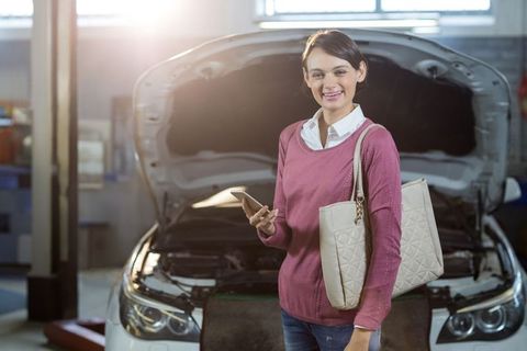 Smiling Woman in Auto Garage with Car Hood Open Holding Smartphone