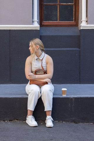 Young woman sitting on city ledge holding leather crossbody purse and takeaway coffee cup