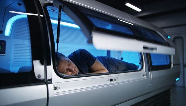 Sleeping adult male reclining inside futuristic metallic sleep capsule with blue lighting