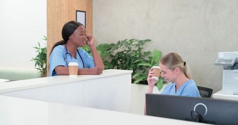 Black and White Nurses Chatting Over Coffee at Clinic Reception Desk During Break
