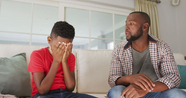 Father looking at son who is visibly upset, sitting on a couch in a modern, bright living room. Showcase emotional support, parent-child relationship, communication skills, family bonds. Suitable for content about parenting advice, emotional well-being, family interactions, and mental health.