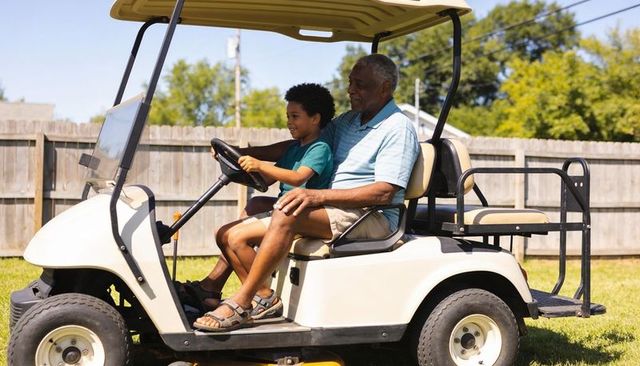 Grandfather and grandson driving white golf cart in sunny backyard, smiling family bonding