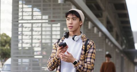 Young male commuter standing on transit platform holding smartphone and wearing headphones