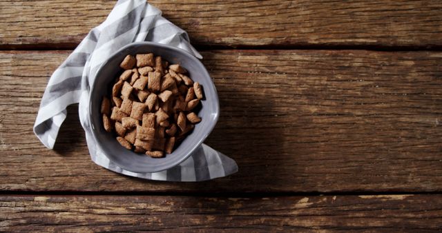 Close-up view of a bowl of chocolate cereal on a rustic wooden table with a striped cloth. Ideal for food blogs, breakfast menus, healthy eating guides, or advertisements for cereals and morning foods. Emphasizes simplicity, authenticity, and a homespun atmosphere, perfect for conveying nutritious and casual meal settings.