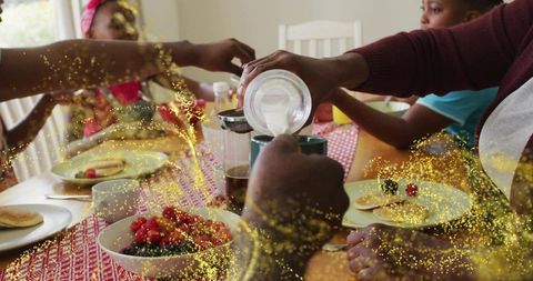 Family pouring milk at sunlit brunch table with pancakes, berries and cozy togetherness
