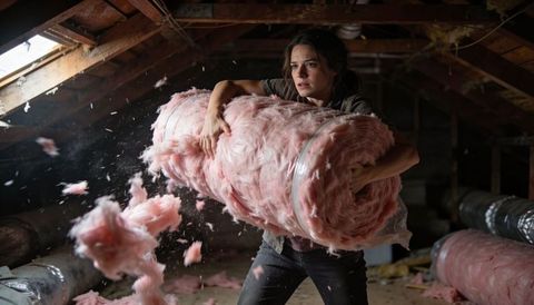 Woman carrying pink fiberglass insulation roll in dusty attic during renovation