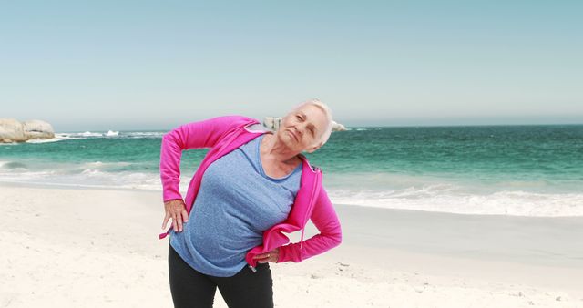 An elderly woman is performing a stretching exercise on a beach with the ocean in the background. This scene is perfect for illustrating senior fitness, healthy lifestyle choices for the elderly, promoting outdoor activities, physical wellness campaigns, and advertisements for senior health and fitness products.