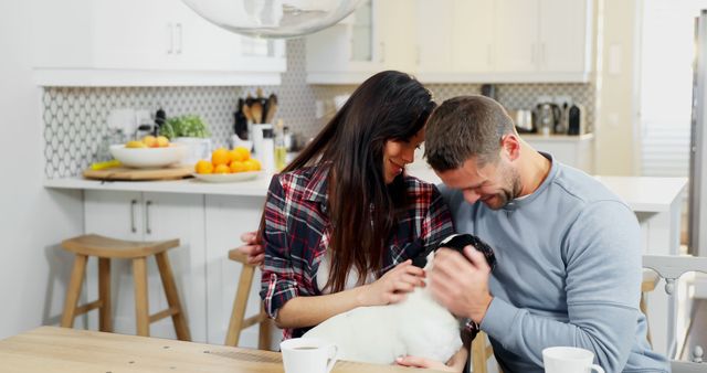 A couple is sitting together at a kitchen table, affectionately cuddling their dog. The kitchen is bright and modern, with various utensils and bowls of fruit visible. This image conveys themes of domesticity, affection, and morning routines. It can be used for websites, blogs, or advertisements related to home life, pet ownership, relationships, and kitchen or home design.