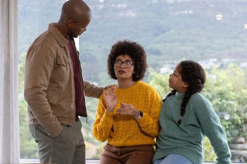 Diverse family moment with father supporting mother while daughter listens on window seat