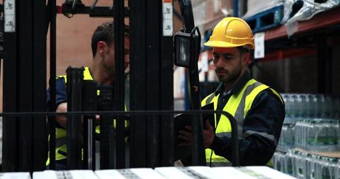 Warehouse workers checking tablet while operating forklift wearing hard hat, hi-vis vests