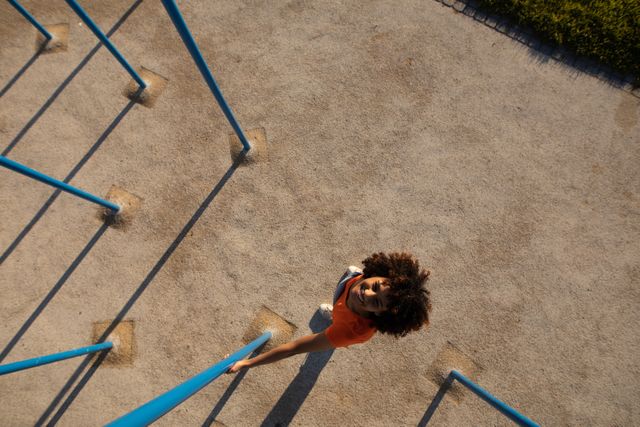 High angle view of a happy biracial woman wearing an orange t shirt hanging out in a children playground on a sunny day looking up to camera and smiling