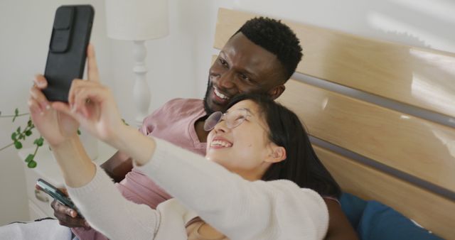 Happy diverse couple taking selfie and lying in bedroom. Spending quality time at home concept.