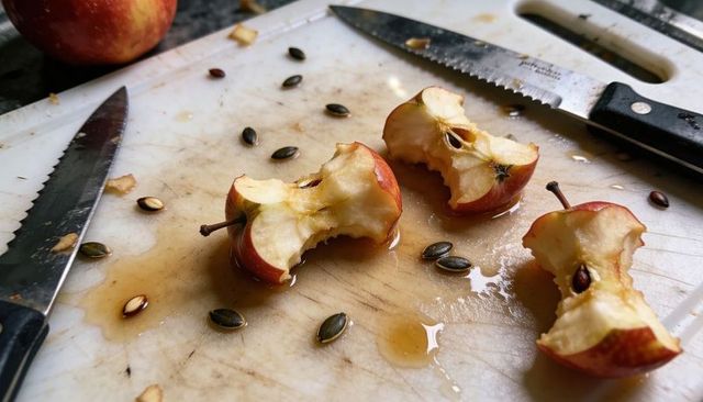 Bitten apple cores dripping juice on white cutting board with serrated knives and seeds