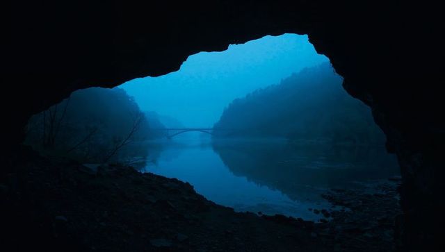 Cave framing misty river valley with arched bridge reflection in blue foggy twilight