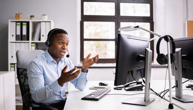 Remote customer support agent gesturing, speaking on headset at dual-monitor workstation