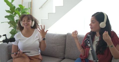 Two women recording podcast on couch in home studio with headphones and microphones