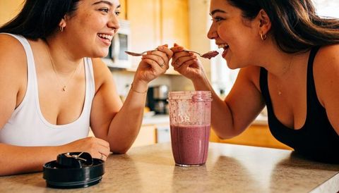 Two women sharing purple smoothie and laughing at kitchen counter, casual wellness moment