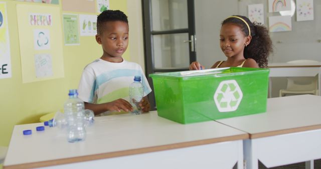 Image of happy african american girl and boy sorting plastic bottles for recycling in classroom. primary school education and learning concept.