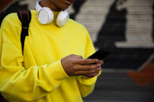 Front view close up of a fashionable biracial transgender in the street, holding and using a smartphone, with headphones on a neck