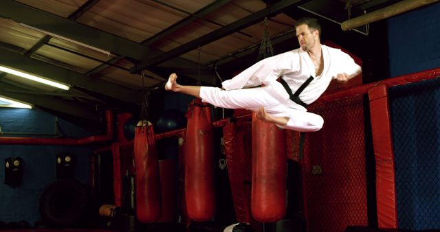 This captures a male martial artist in white karate uniform with black belt executing a high flying kick with precision and strength at a gym. Surroundings include heavy punching bags and training equipment, emphasizing the intense training atmosphere. Ideal for use in advertisements for martial arts schools, fitness classes, motivational posters, and action-sequence illustrations.