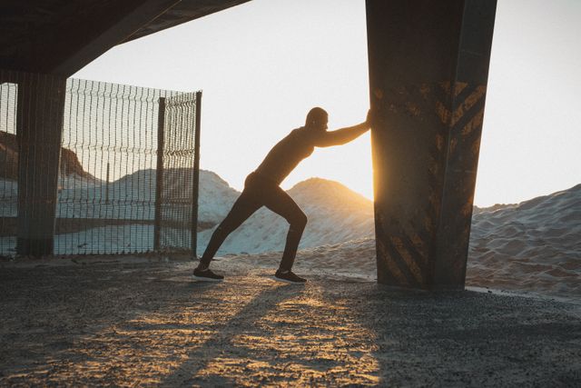 African american man exercising, stretching under bridge on beach at sunset. healthy outdoor lifestyle fitness training.