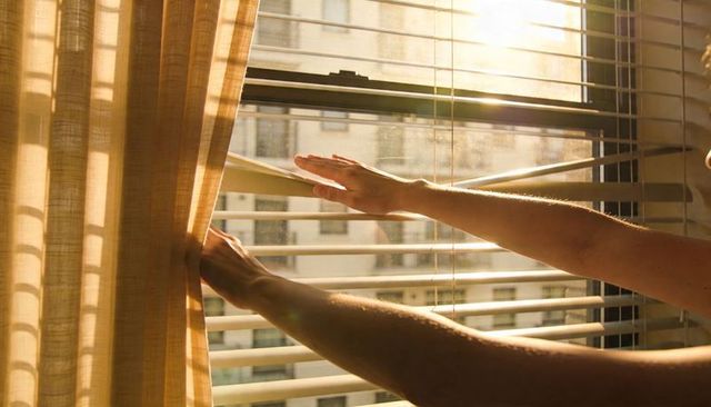 Morning sunlight through blinds as hands open curtain and adjust venetian shades