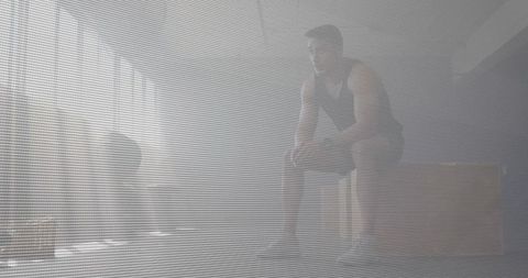 Male athlete sitting on plyo box resting in hazy sunlit gym with blinds and texture