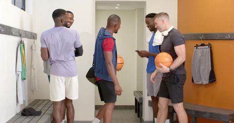 Multiracial basketball teammates chatting in locker room with balls and phone after workout