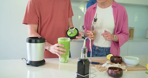 Couple preparing green smoothie at kitchen island pouring oil and blending ingredients