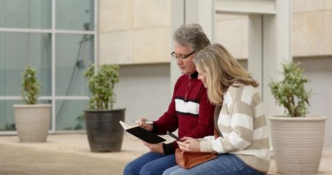 Retired couple planning trip together using notebook and smartphone on urban bench