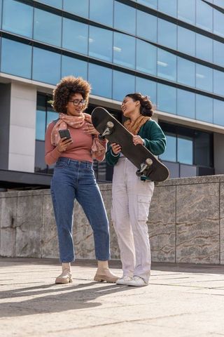 Diverse young women sharing skateboard and smartphone in urban plaza casual autumn style