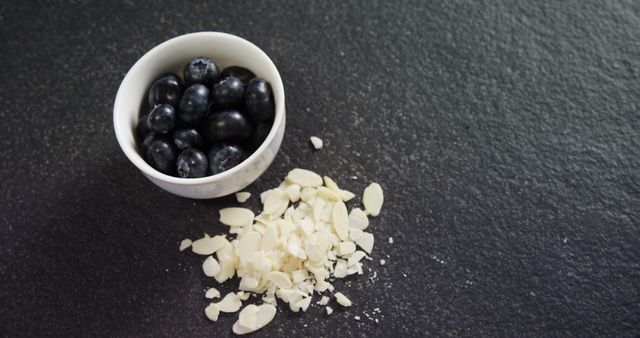Image shows white bowl filled with blueberries next to scattered sliced almonds on dark surface. Ideal for promoting healthy eating, showcasing nutritious snack options, or adding visuals to food blogs and wellness articles.