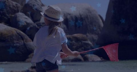 Child wading with red net on rocky coast wearing straw hat and shorts, sunlit tidepool