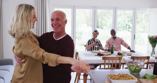 This vibrant photo shows two senior couples celebrating in the dining room at home. One couple is dancing in the foreground, exhibiting joy and happiness, while the other couple is seated at the table, smiling and engaging in conversation. The table is beautifully set with various food dishes, including a casserole and a salad. This cheerful and diverse scene promotes themes of rediscovery in later life, togetherness, and friendship. Perfect for advertisements targeting retirement activities, social engagements, or family lifestyle content.