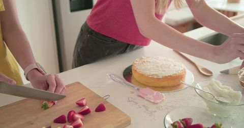 Slicing strawberries and decorating sponge cake on sunny home kitchen countertop