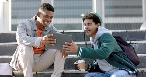 Young diverse men sitting on campus steps sharing tablet and coffee, urban students
