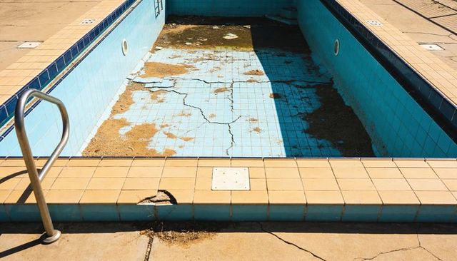 Drained blue tiled pool showing large cracks, sediment patches and metal ladder on deck