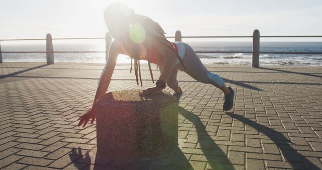 Woman with long braids practicing a challenging outdoor exercise routine on a pier with ocean in the background. Bright sunlight creating lens flare. Themes of health, wellness, fitness, and dedication. Useful for fitness training, health blogs, motivational content, and exercise tutorials.