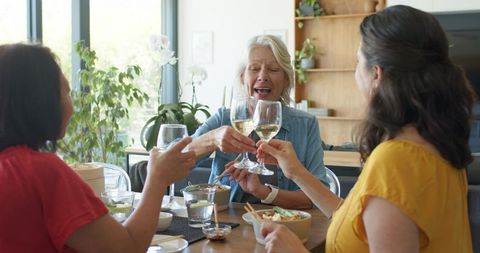 Diverse women toasting white wine while sharing casual home lunch and laughing