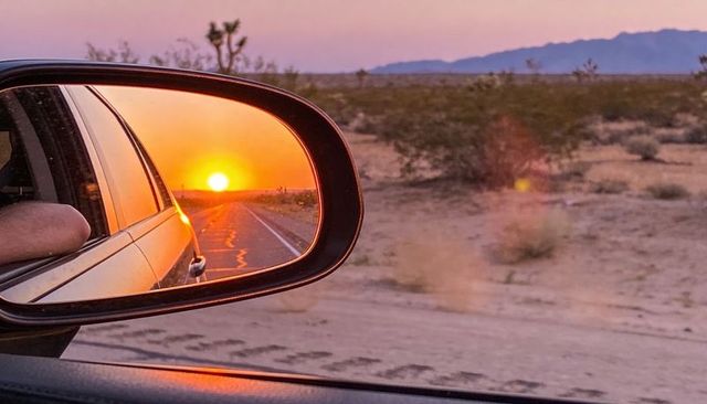Sunset reflected in car side mirror on desert highway, arm resting out window at dusk