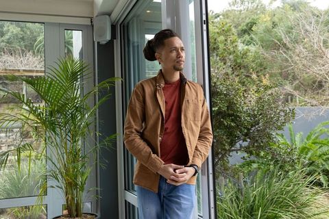 Asian man standing in glass doorway looking out at lush garden wearing wristwatch
