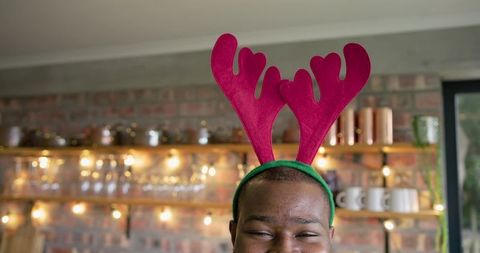 African american man wearing magenta reindeer antlers smiling in cozy cafe with lights