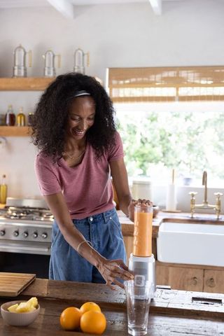 African american woman making fresh orange smoothie in sunlit rustic farmhouse kitchen