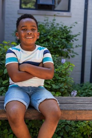 African american boy smiling on wooden bench in garden in striped tee and denim shorts