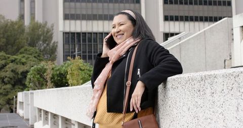 Mature woman smiling while talking on smartphone, leaning on concrete railing in city plaza