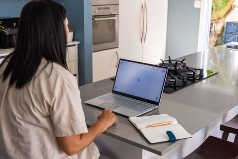 Mature woman working at kitchen island with laptop and notebook, minimalist home workspace
