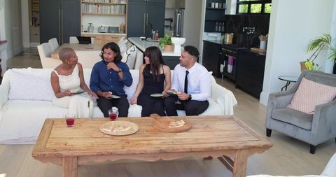 African american friends sitting on white sofa sharing pizza in sunlit open-plan kitchen