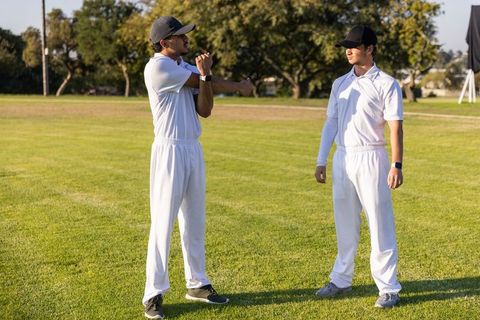 Diverse cricket teammates stretching and standing during sunlit warmup on grass field