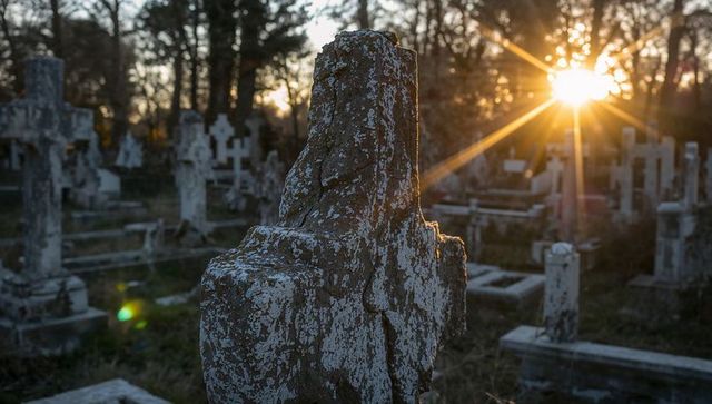 Lichen-covered stone cross at sunset in weathered cemetery with sunburst lens flare