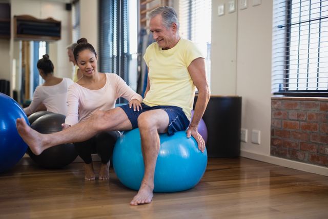 Female therapist helping senior male patient doing leg exercise on blue ball at hospital ward